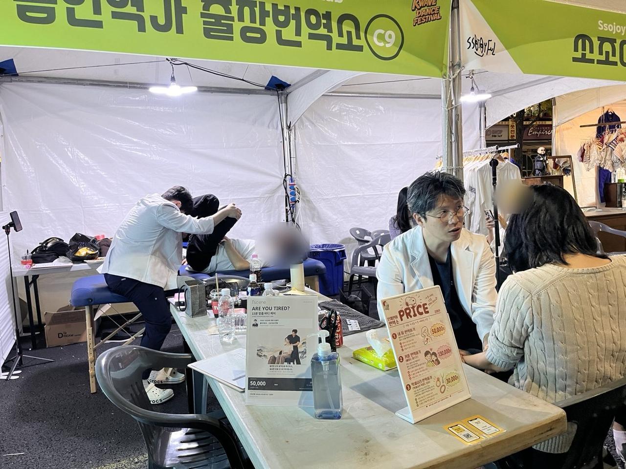 축제 부스에서 상담 테이블에 앉아 게스트의 이야기를 듣는 김윤하와, 뒤쪽 테이블에서 진행 중인 케어